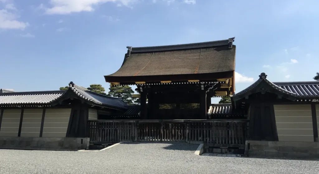 A traditional wooden gate with a curved, thatched-style roof at the Kyoto Imperial Palace, flanked by low tiled-roof walls, set on a gravel courtyard under a blue sky with scattered clouds. A traditional wooden gate with a curved, thatched-style roof at the Kyoto Imperial Palace, flanked by low tiled-roof walls, set on a gravel courtyard under a blue sky with scattered clouds.