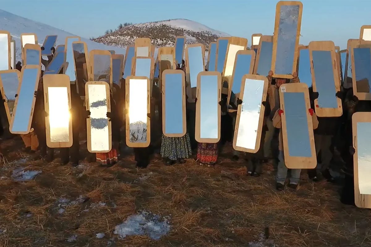 Dozens of Indigenous water protectors hold large mirrored shields during the Standing Rock protests. The reflective surfaces face forward, creating a striking visual barrier that mirrors the surrounding landscape and people.