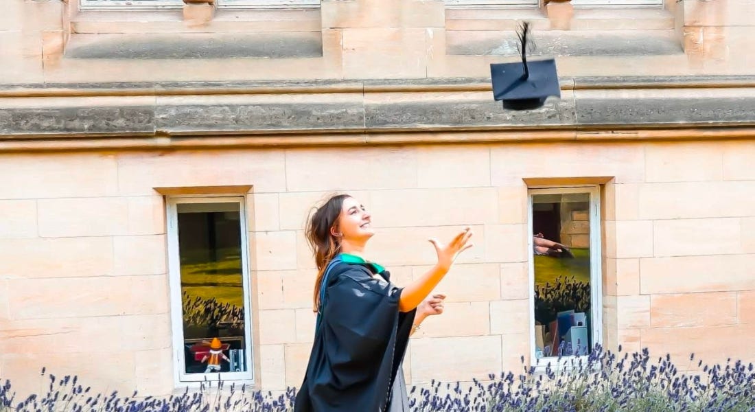 A woman - Eve Smallman - throwing her graduation cap in the air with a stone building behind her