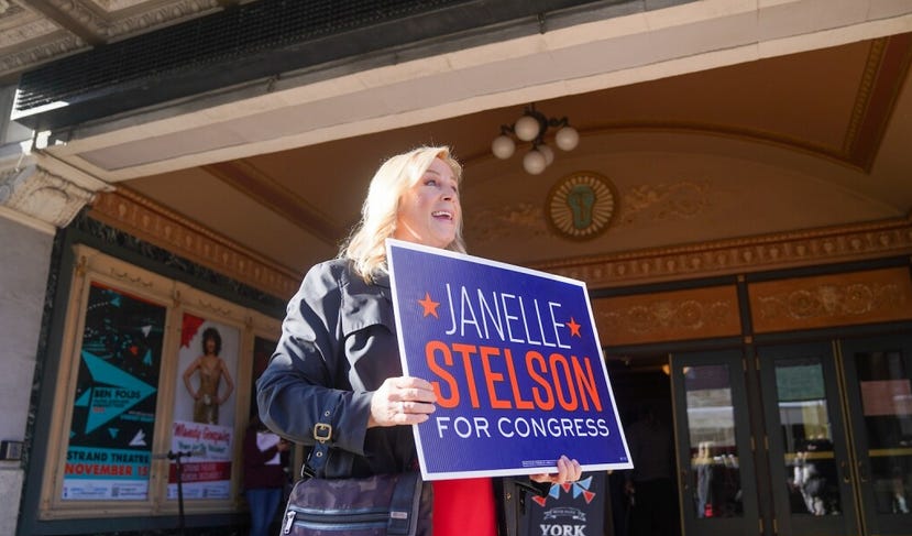 A blonde woman in a jean jacket standing in the entryway to a movie theater holds up a sign reading "Janelle Stelson for Congress."