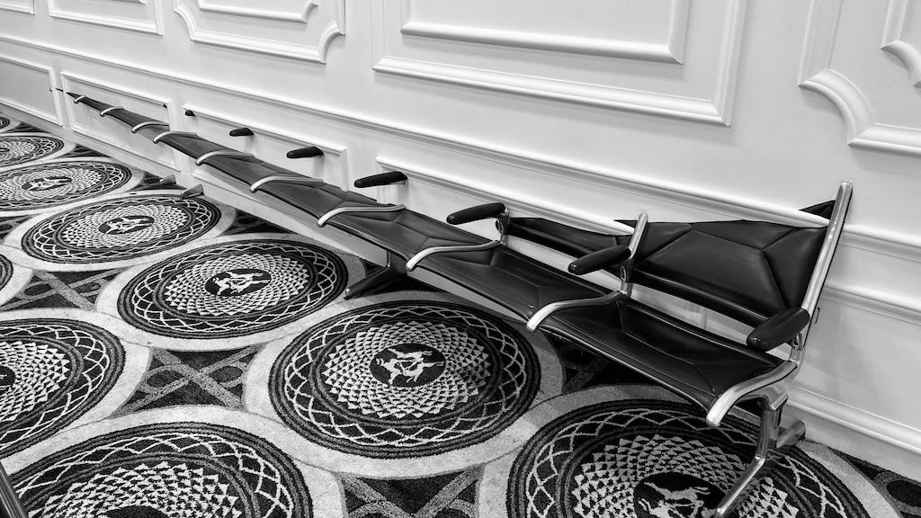 In this black-and-white photo, a long row of connected metal-framed chairs is installed so that each seat appears to slice seamlessly through the white paneled wall, as if passing in and out of it. Only partial sections of the chairs are visible on each side of the wall surface, creating a surreal, impossible bench. The chairs rest over patterned carpet with bold circular geometric designs. In this black-and-white photo, a long row of connected metal-framed chairs is installed so that each seat appears to slice seamlessly through the white paneled wall, as if passing in and out of it. Only partial sections of the chairs are visible on each side of the wall surface, creating a surreal, impossible bench. The chairs rest over patterned carpet with bold circular geometric designs.