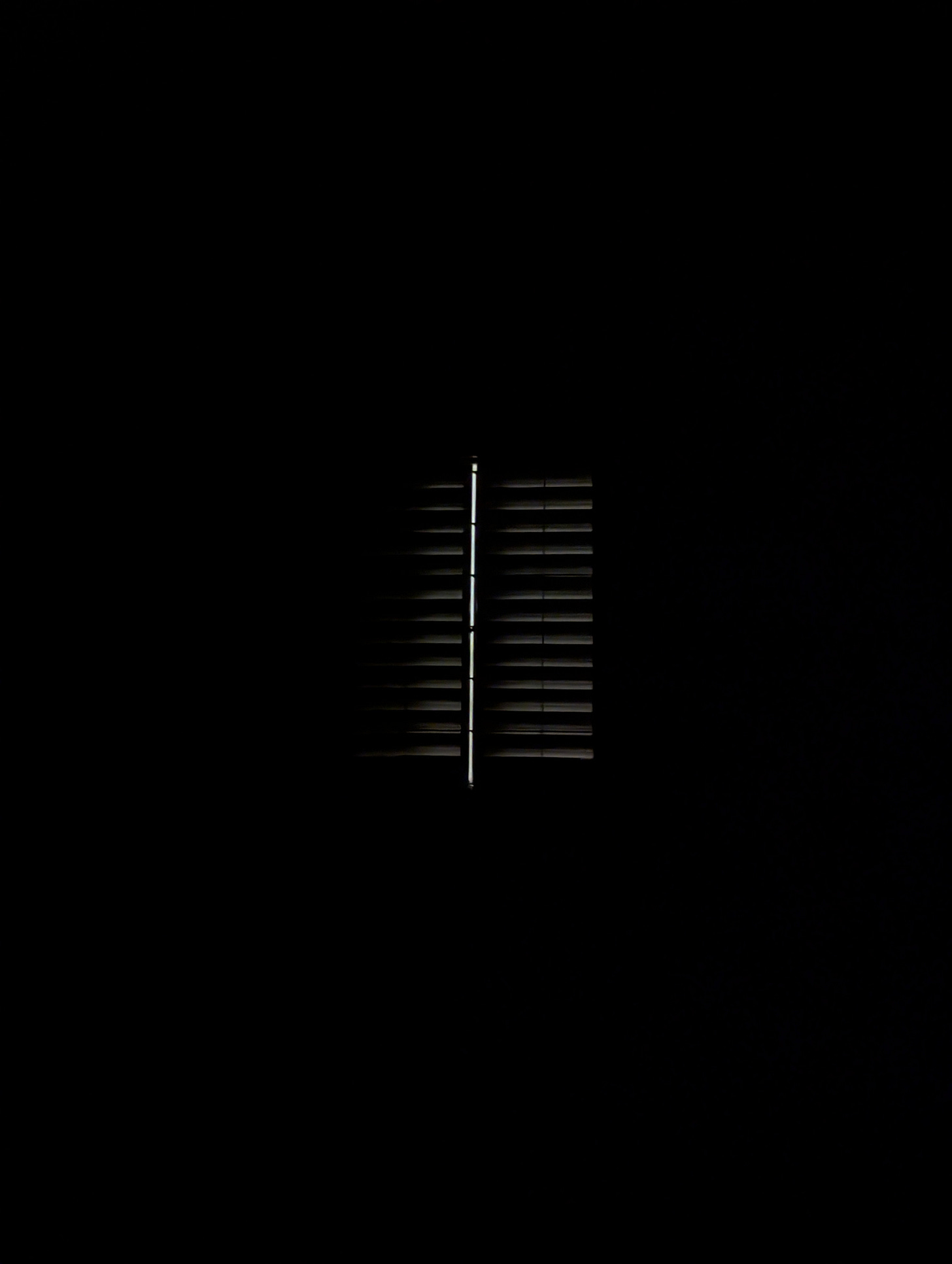 A single illuminated window with horizontal blinds glows against complete darkness, creating a stark contrast between the warm light filtering through the slats and the surrounding black void. A single illuminated window with horizontal blinds glows against complete darkness, creating a stark contrast between the warm light filtering through the slats and the surrounding black void.