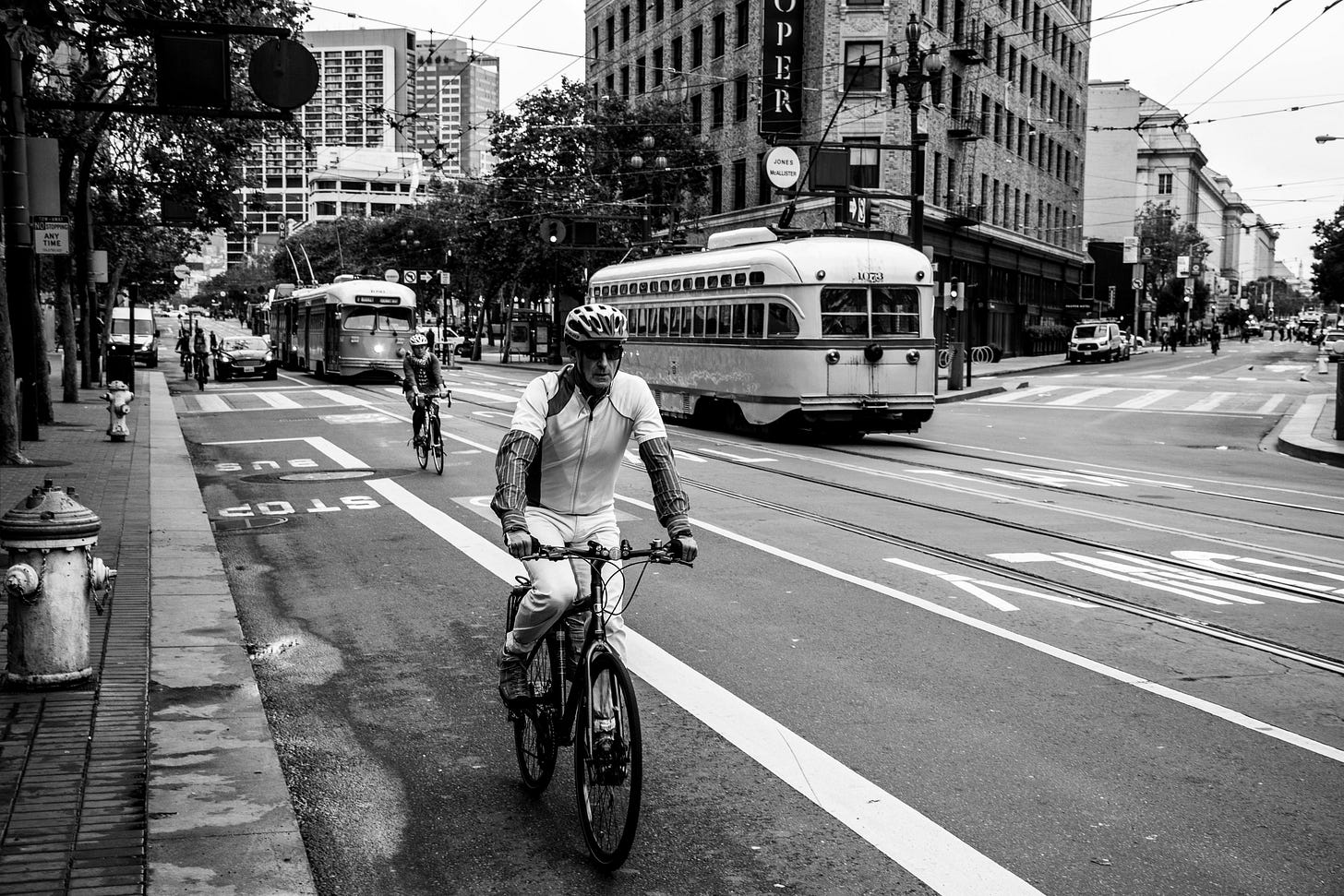 A black and white photo of a man cycling down Market Street in San Francisco with trams behind him