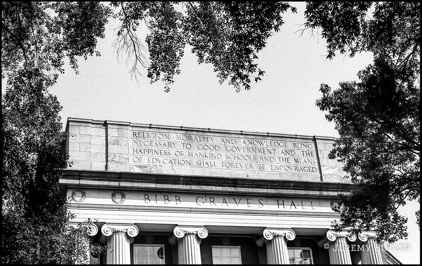 Black and white photo of Bibb Graves Hall at the University of Alabama facade with Ionic columns and carved stone inscription framed by trees.