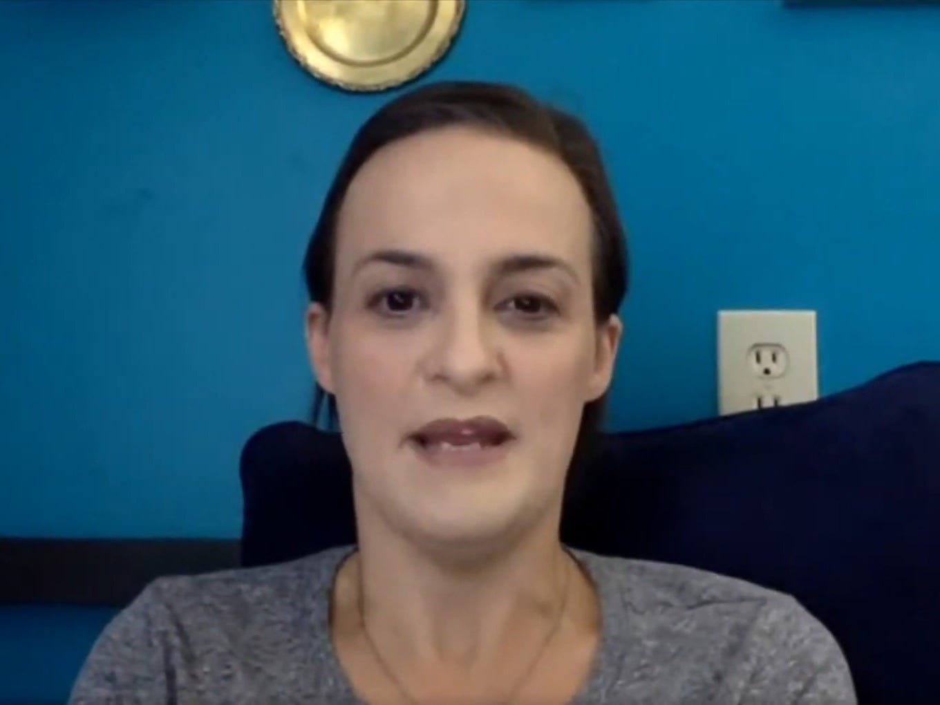 Elisabeth Finch wearing a gray shirt and sitting in a chair with a blue wall behind her.