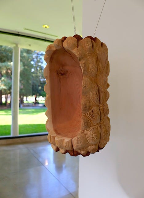 Hollowed redwood sculpture suspended by thin steel wires, with a soft pink interior and a carved exterior that reveals layered wood grain and rounded contours. Hollowed redwood sculpture suspended by thin steel wires, with a soft pink interior and a carved exterior that reveals layered wood grain and rounded contours.