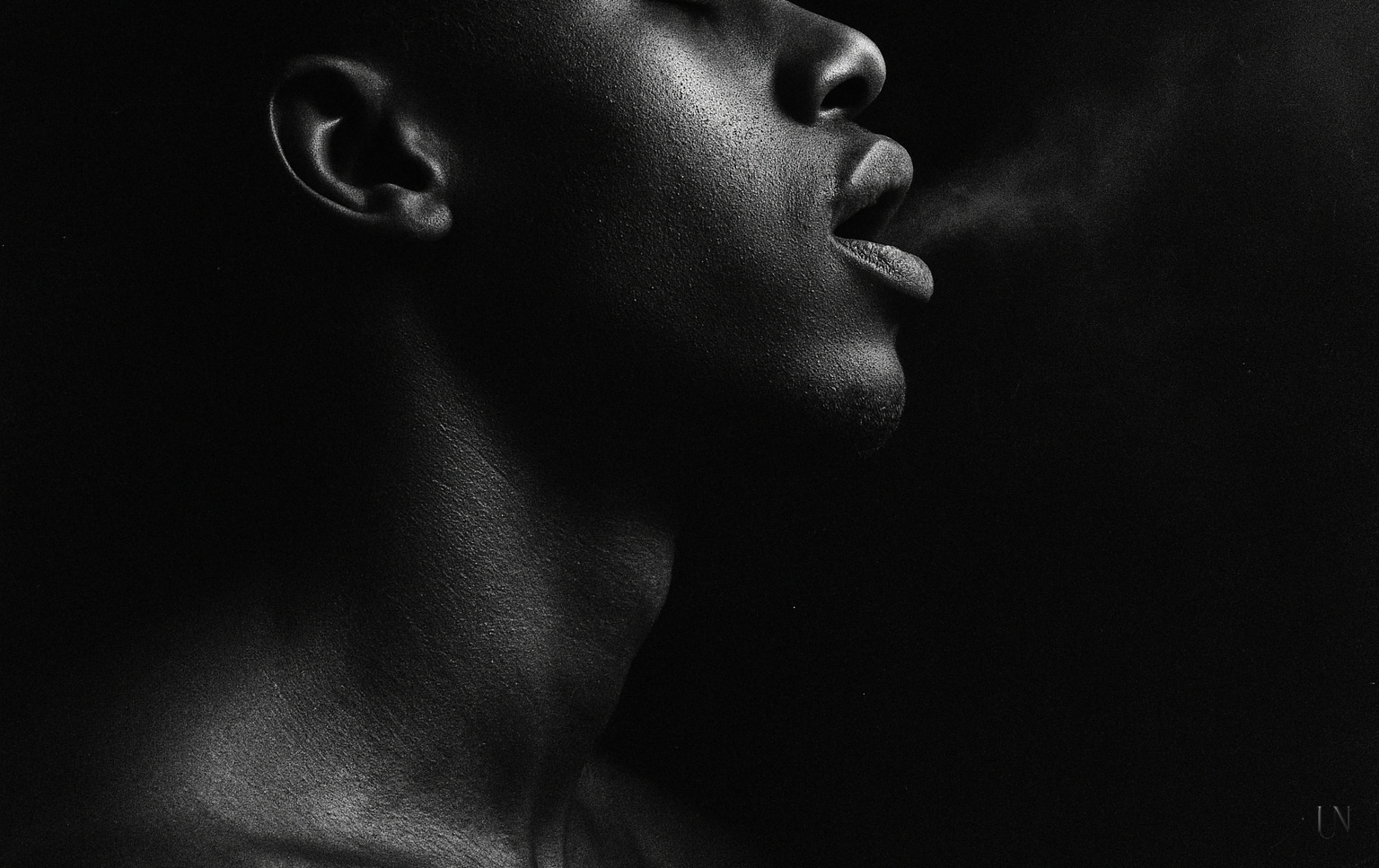 A Black male figure in close profile, eyes closed, lips parted mid-breath. A faint exhale leaves his mouth like smoke, caught in low sidelight. His throat and jaw glimmer softly while the rest dissolves into blackness—an image of tenderness reclaiming breath.