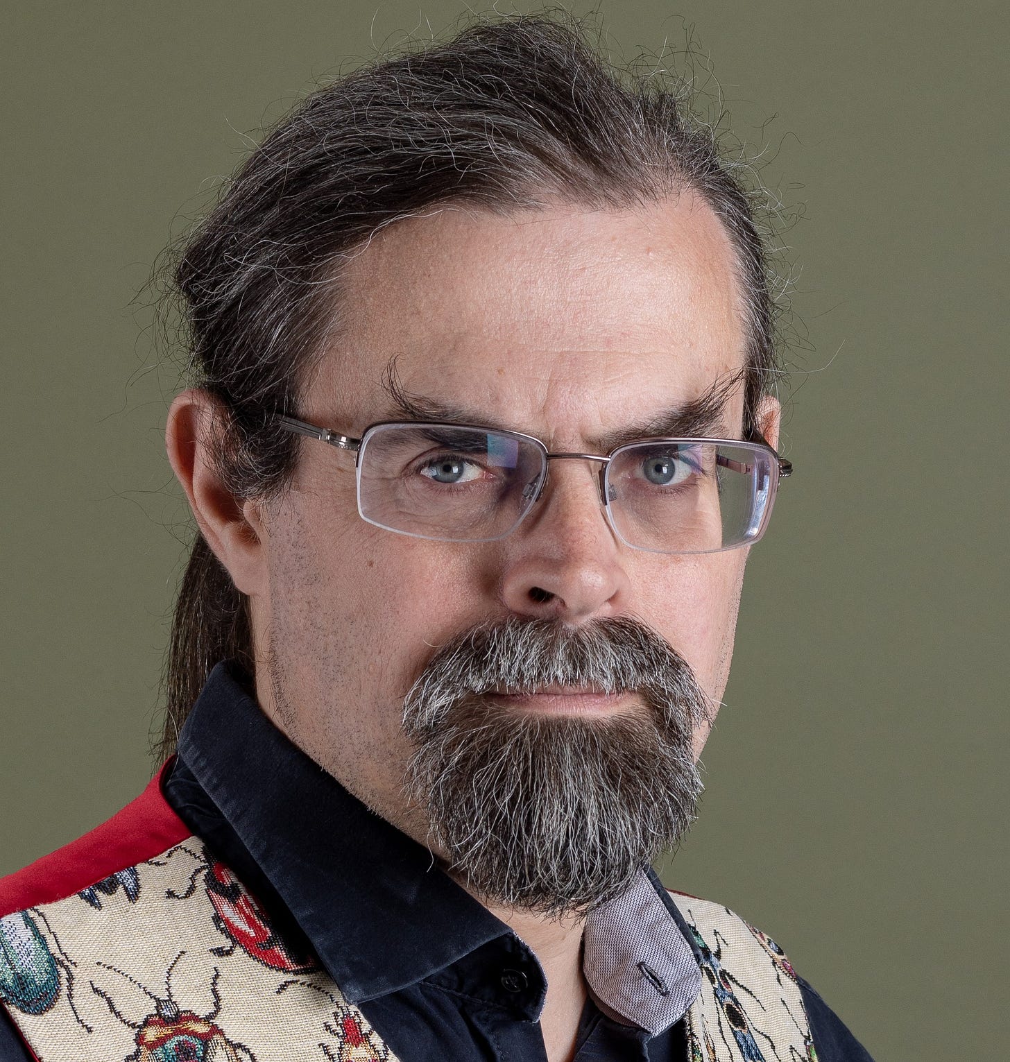 Studio portrait of a man with glasses, tied-back dark hair, and a grey beard, wearing a black shirt and a cream waistcoat patterned with colourful beetles. Studio portrait of a man with glasses, tied-back dark hair, and a grey beard, wearing a black shirt and a cream waistcoat patterned with colourful beetles.