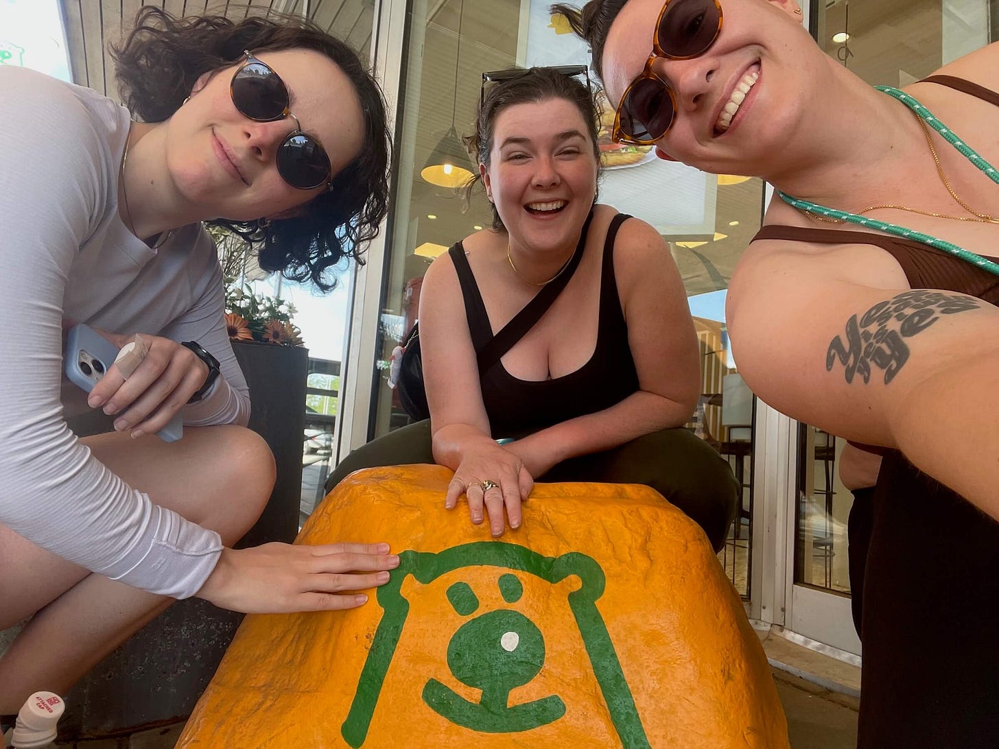 Elizabeth, Jackie, and Shell all lean in around an orange rock with a happy green bear painted on it. Elizabeth, Jackie, and Shell all lean in around an orange rock with a happy green bear painted on it.