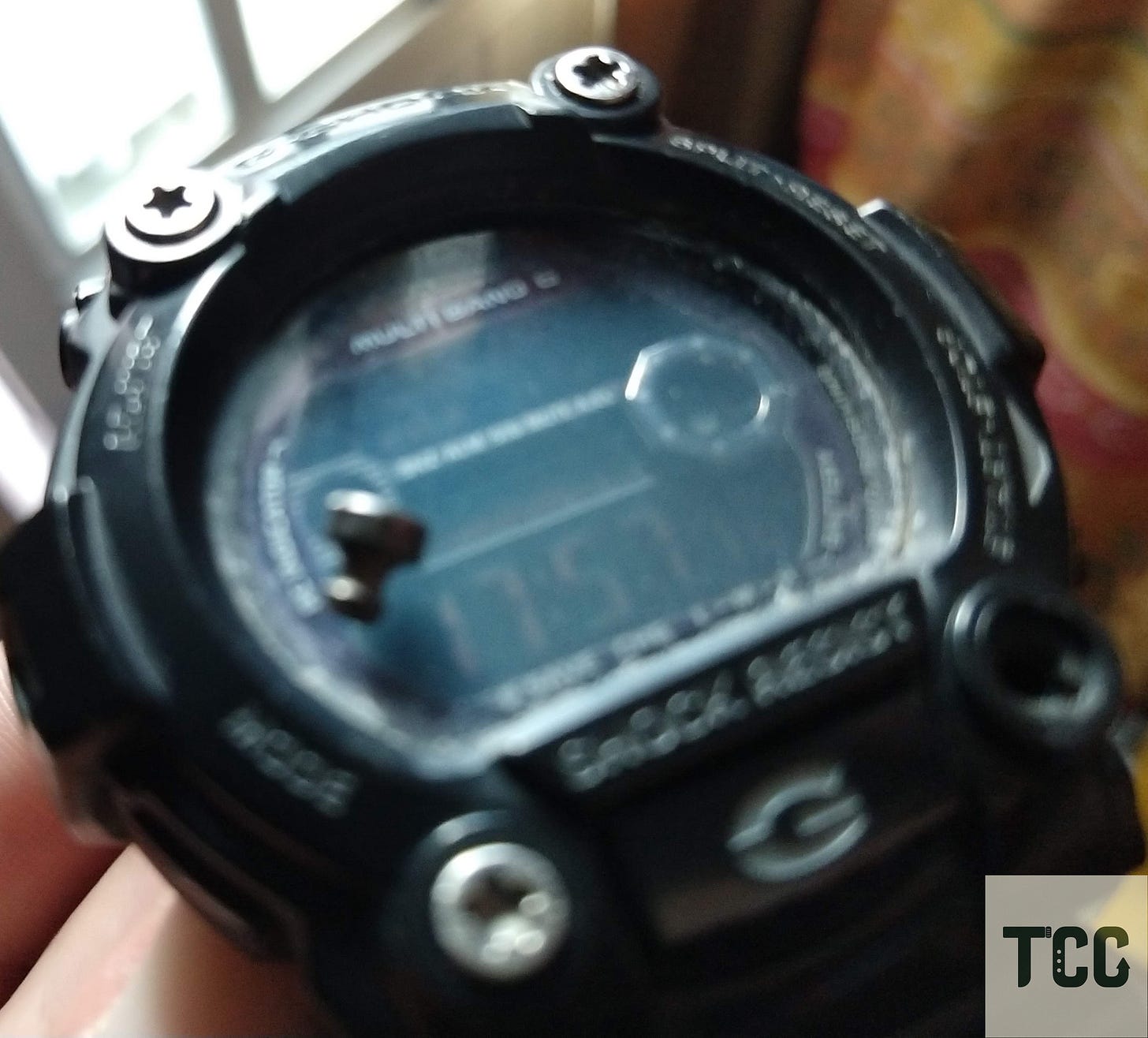 Close-up, slightly blurry photo of a black G-Shock digital watch with a damaged screen. A loose metallic piece sits on the glass, and the watch appears scratched and worn.
