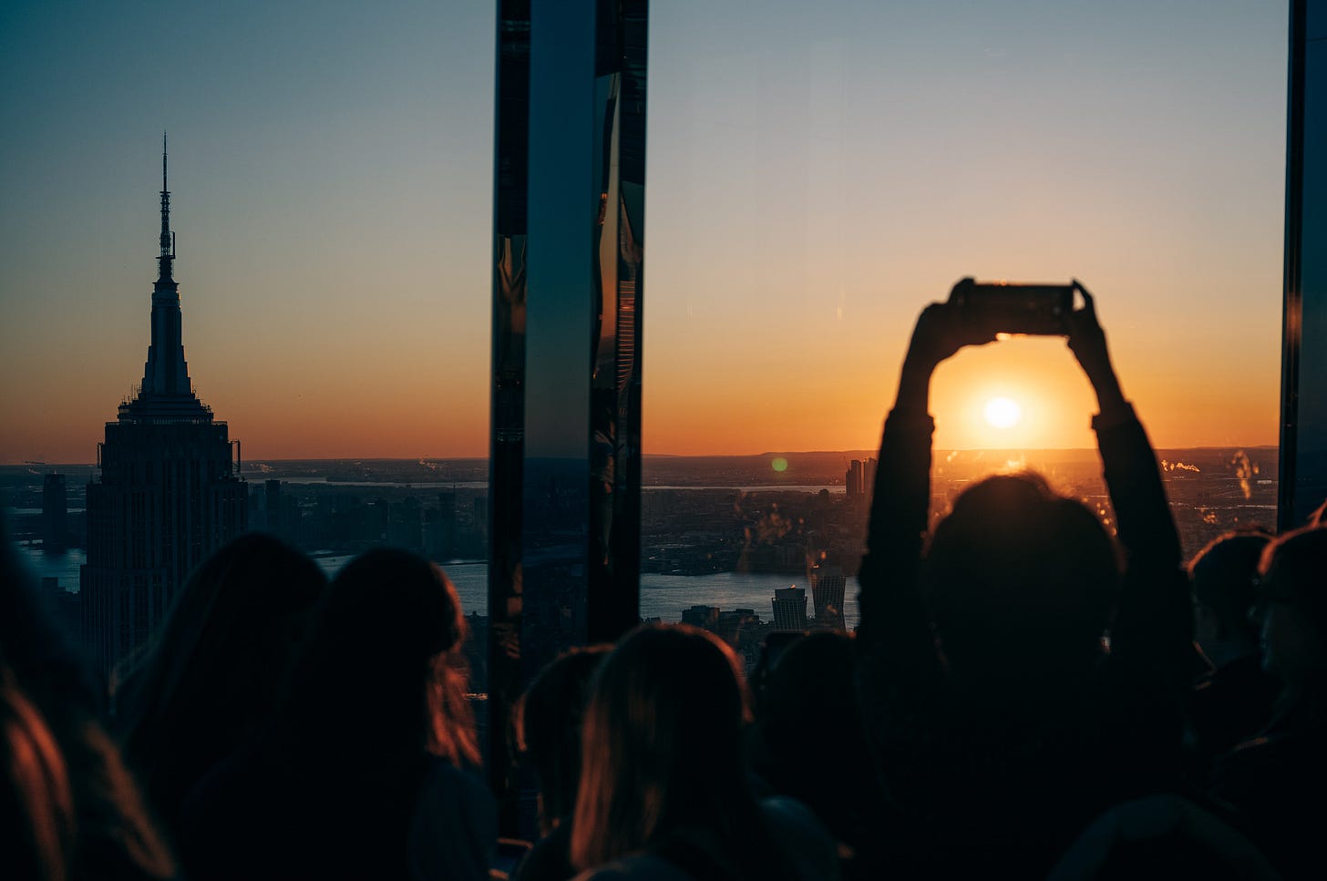 A crowd silhouetted at the Summit One Vanderbilt observation deck at sunset, one person holding a phone up so the setting sun sits inside the screen, with the Empire State Building at left, New York.