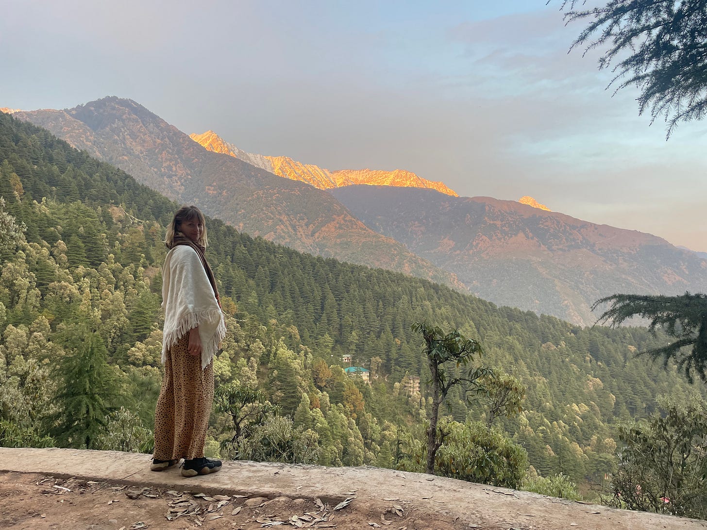 Faith is standing on a ledge in front of a mountainscape with pine trees in the foreground and mountains in the background. The further peaks are snowy. Faith is standing on a ledge in front of a mountainscape with pine trees in the foreground and mountains in the background. The further peaks are snowy.