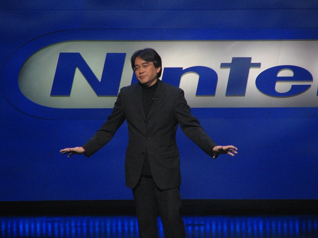 A Japanese man in a black business suit stands on stage with his arms spread outward some. Behind him is a blue wall with a blue Nintendo logo partially in view. A Japanese man in a black business suit stands on stage with his arms spread outward some. Behind him is a blue wall with a blue Nintendo logo partially in view.