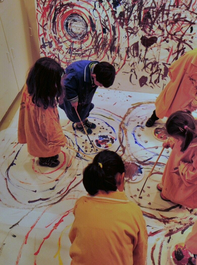 a yellowed photo of 5 children in a room painting on a large canvas with sticks, having completed many colorful concentric circles