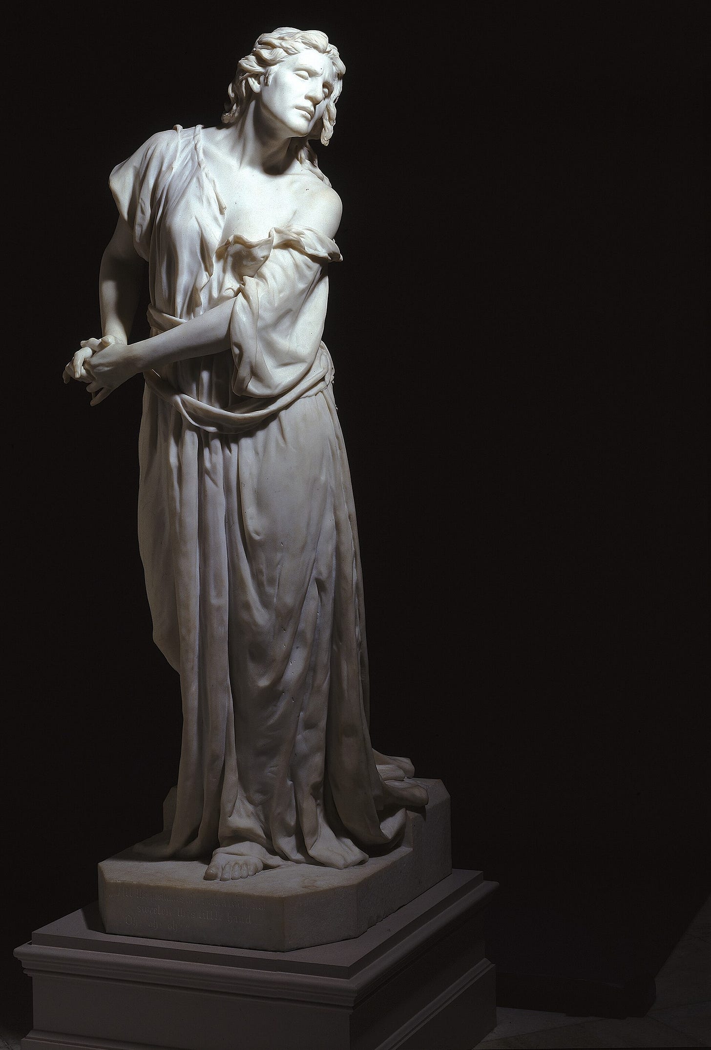 A life-size white marble woman in a long, sharply folded gown walks forward with her eyes half closed, her brow furrowed and one arm held stiffly at her side. A life-size white marble woman in a long, sharply folded gown walks forward with her eyes half closed, her brow furrowed and one arm held stiffly at her side.