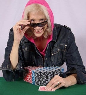 Photo of of Author Donna Blevins in 2009 sitting at a poker table with green felt, with stacks of black, blue and red chips in front of her. She has on a dark jean jacket, a hot pink hoodie over her had, and she is looking over sun glasses. With her left hand she is protecting her hole cards.