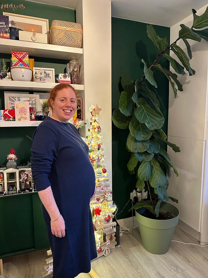 A pregnant woman (& her cat) in front of a wooden Christmas tree and some shelves