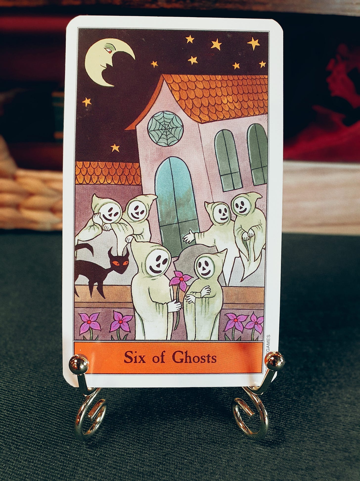 Six of Ghosts: The Six of Ghosts tarot card from Kipling West’s Halloween Tarot, showing six hooded ghosts outside a building at night, with one ghost offering another a pink flower beneath a crescent moon and stars. Six of Ghosts: The Six of Ghosts tarot card from Kipling West’s Halloween Tarot, showing six hooded ghosts outside a building at night, with one ghost offering another a pink flower beneath a crescent moon and stars.