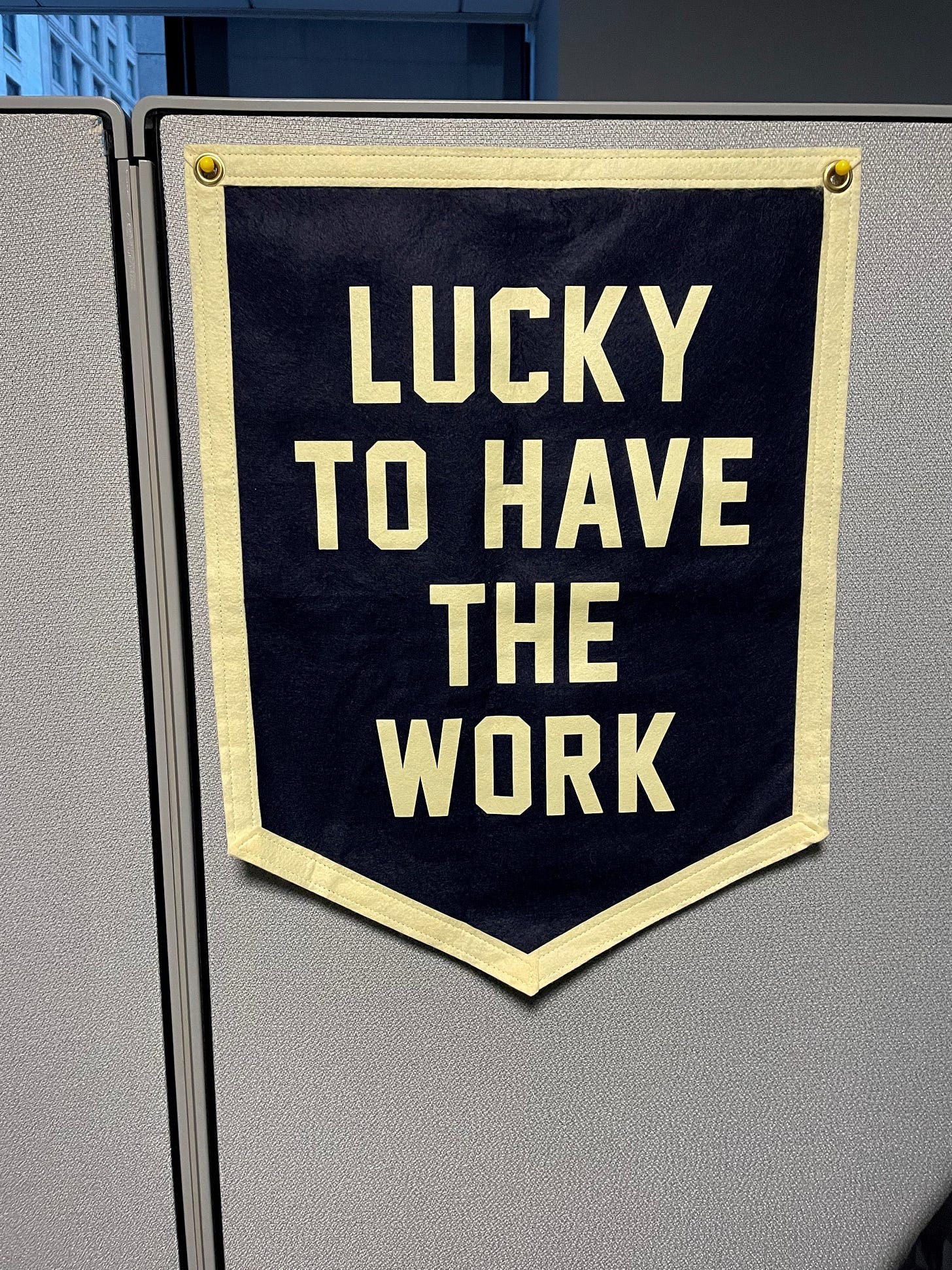 A banner on a cubicle wall. The banner has cream edging and the center of the banner is a blue felt field. On the blue field in white letters the banner reads 'Lucky To Have the Work.'