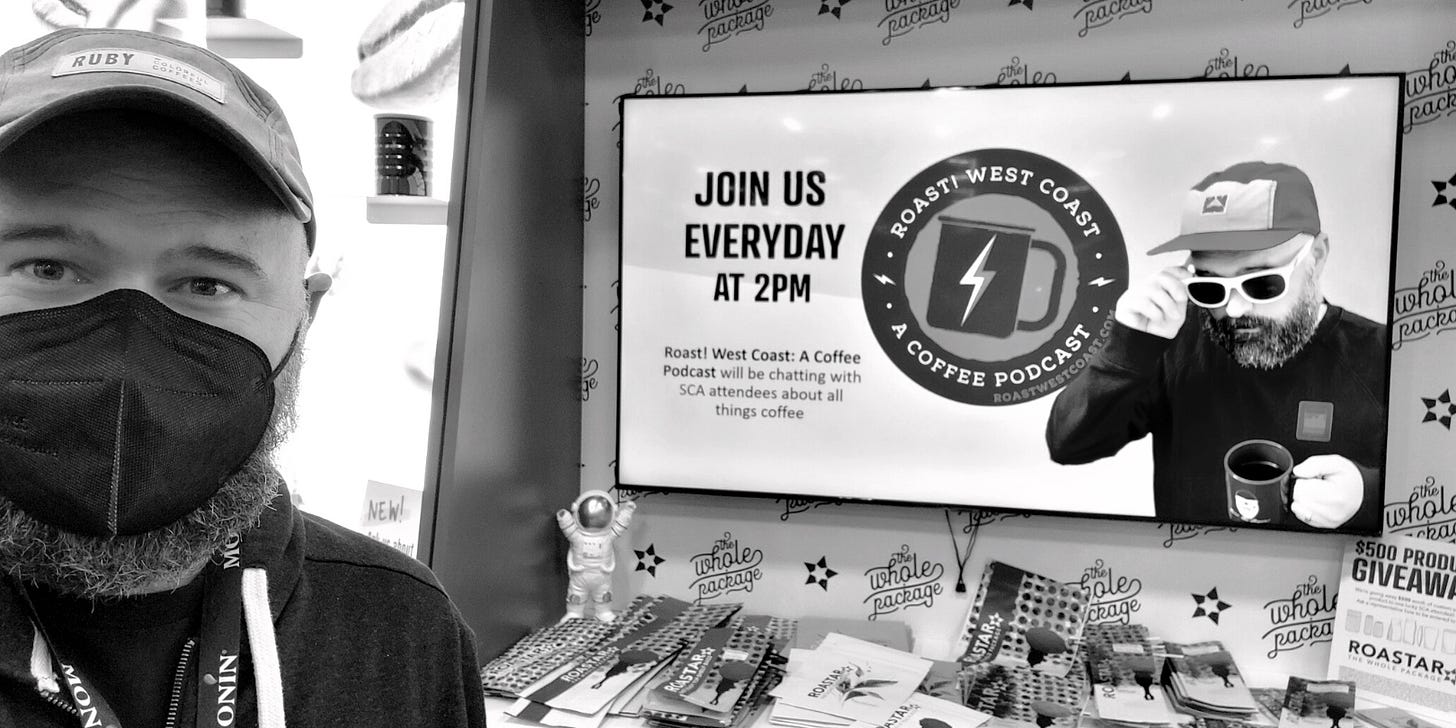 A bearded, masked (n95) man wearing a ball cap looks at the camera while standing next to a sign inviting you to share a cup of coffee with him and a picture of him with out the mask at a convention center booth. Black and white photo.