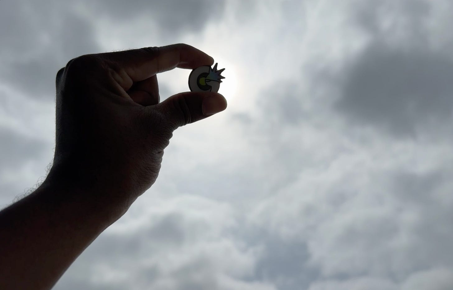 ArmaniXR holds the Glitches.VR pin IRL up against a mostly cloudy sky.