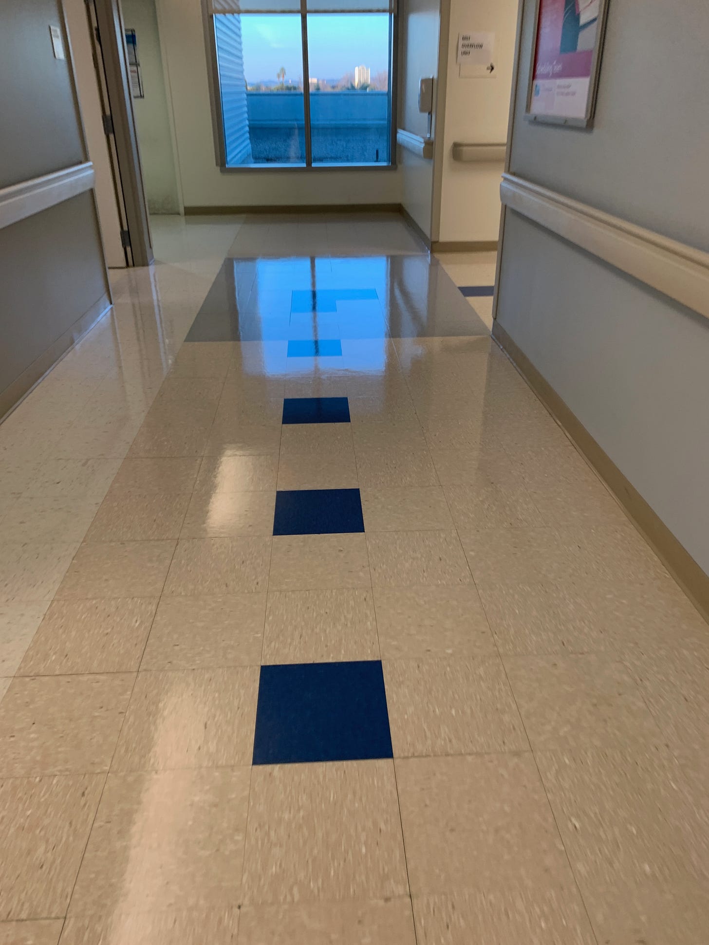 A hospital hallway of white vinyl tiles except and a row of blue tiles in the middle, leading to another hallway.