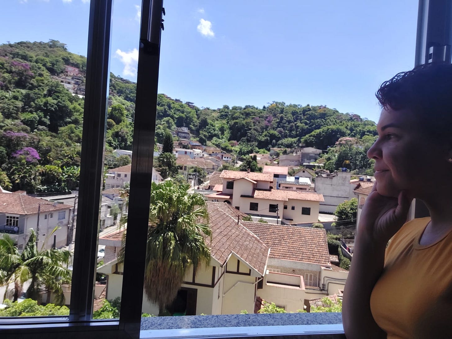 Clara, uma mulher branca de 43 anos e de cabelos curtos olha por uma janela. Lá fora há um dia iluminado, montanhas verdejantes com alguns ipês roxos salpicados e muitas casinhas. Clara, uma mulher branca de 43 anos e de cabelos curtos olha por uma janela. Lá fora há um dia iluminado, montanhas verdejantes com alguns ipês roxos salpicados e muitas casinhas.