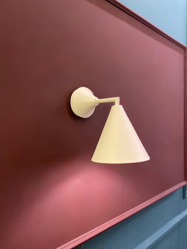 May include: A white wall-mounted light fixture with a cone-shaped shade. The light is mounted on a burgundy wall. May include: A white wall-mounted light fixture with a cone-shaped shade. The light is mounted on a burgundy wall.