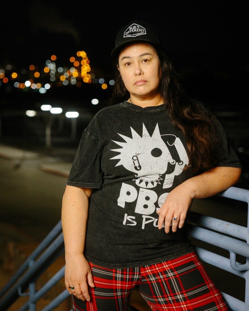 A person — wearing a black hat, black shirt that reads “PBS is punk” and red flannel pants — stands at the top of a flight of blue stairs as they rest their left elbow on the rail. Blurred lights from a nearby refinery can be seen in the background.