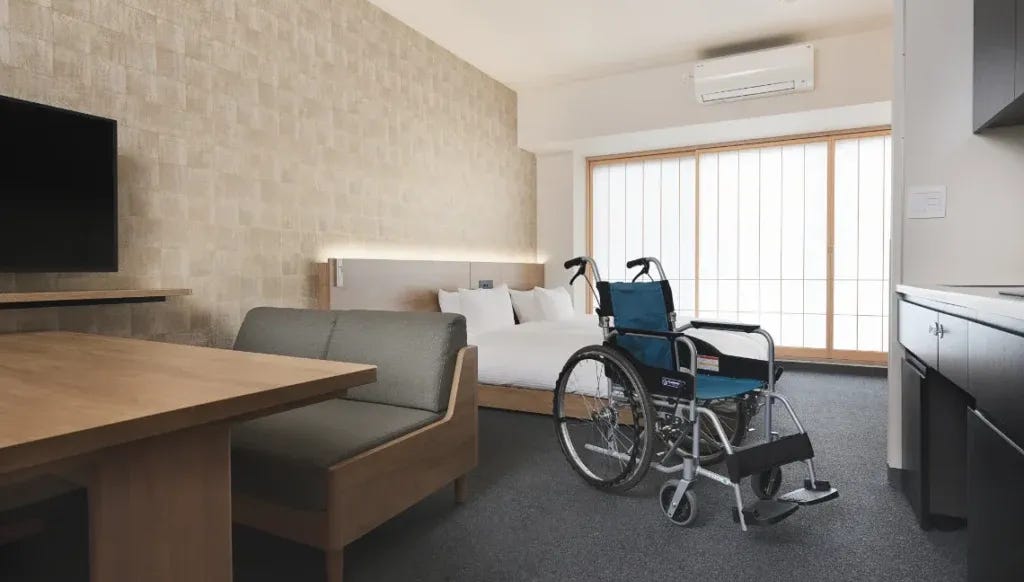 A wheelchair-accessible hotel room at MIMARU Tokyo Kinshicho with a manual wheelchair parked beside the bed, wide open floor space, a kitchenette, dining table, and sliding shoji-style windows leading to a balcony. A wheelchair-accessible hotel room at MIMARU Tokyo Kinshicho with a manual wheelchair parked beside the bed, wide open floor space, a kitchenette, dining table, and sliding shoji-style windows leading to a balcony.