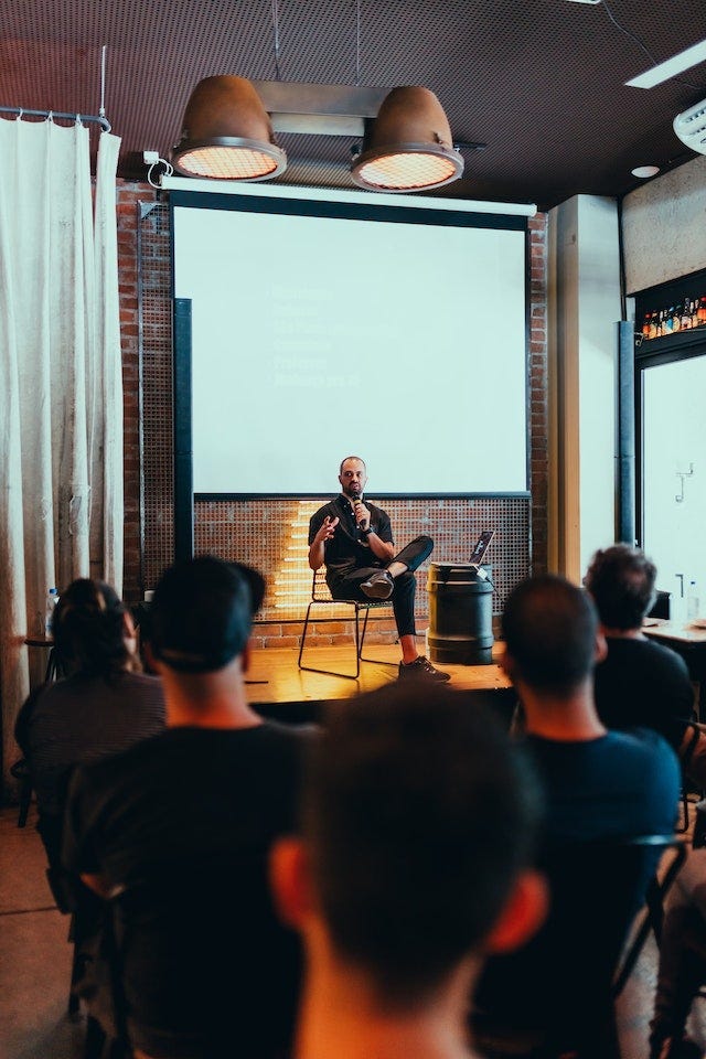 A man sitting in front of a stage, sitting down and speaking into a microphone, There is a captive audience sitting in casual clothes looking at him. A man sitting in front of a stage, sitting down and speaking into a microphone, There is a captive audience sitting in casual clothes looking at him.