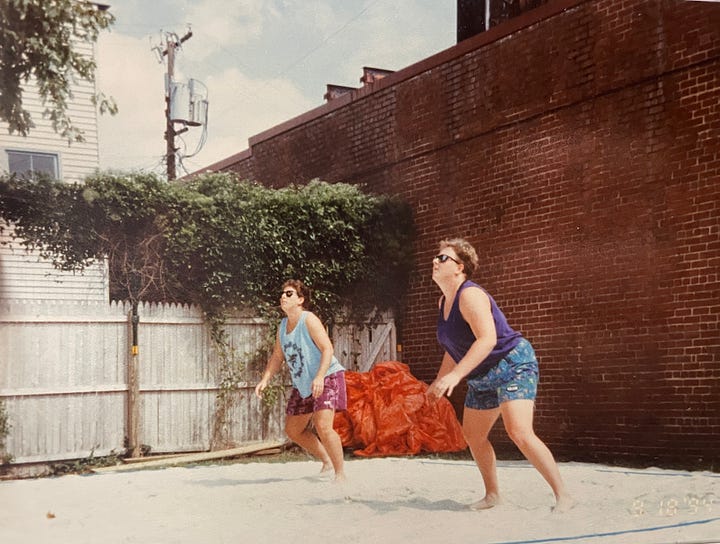 two photos of the same sand volleyball court with brick wall on right of photo and wooden picket fence along background; photo on left shows a platform deck with tables; photo on right shows to women looking up to watch for the ball