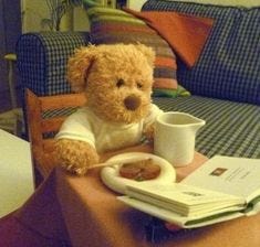 This may contain: a teddy bear sitting at a table with a cup of coffee and book on it