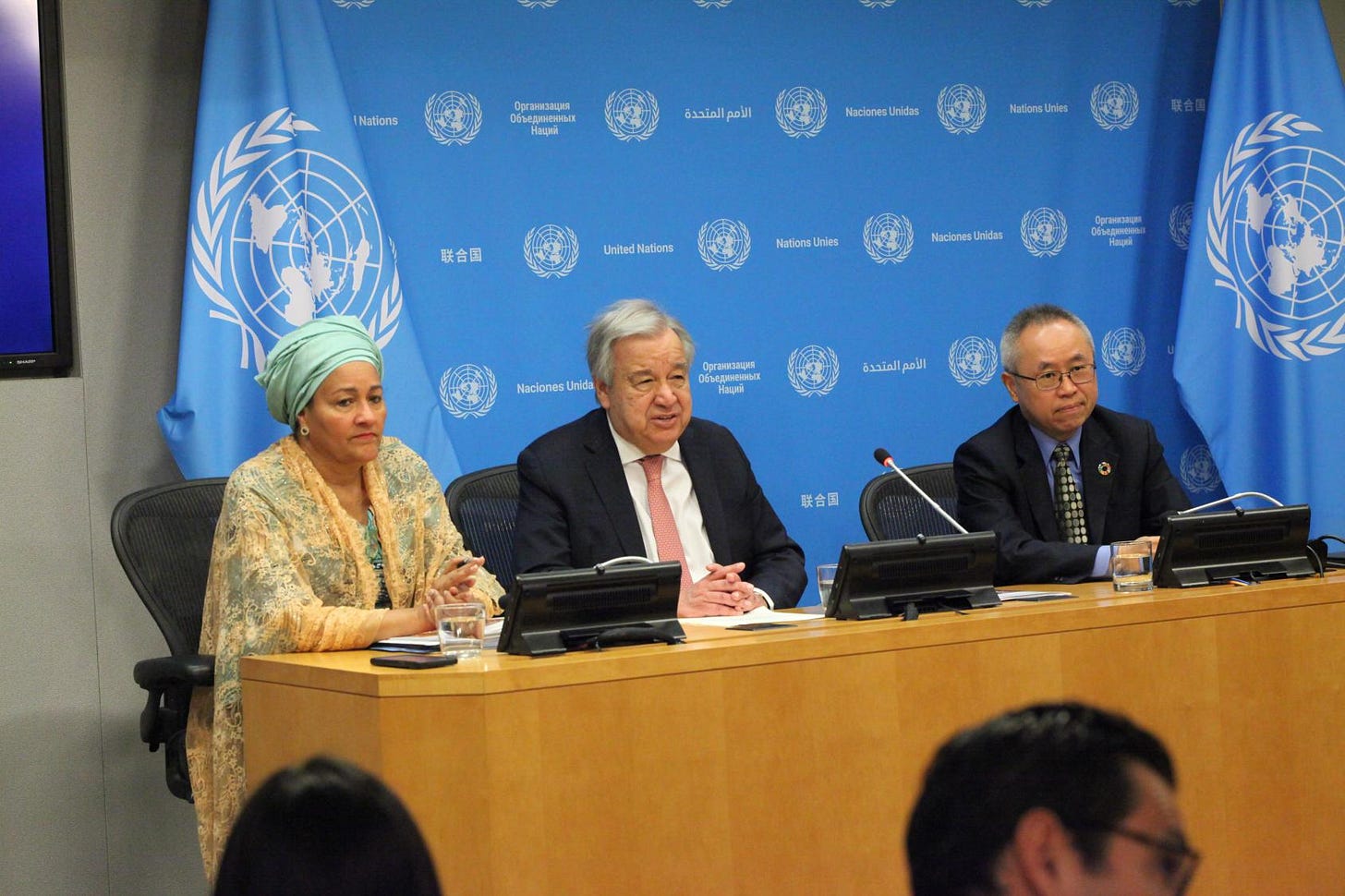 A woman and two men seated at a conference room table. UN flags besides them and a UN backdrop behind them A woman and two men seated at a conference room table. UN flags besides them and a UN backdrop behind them