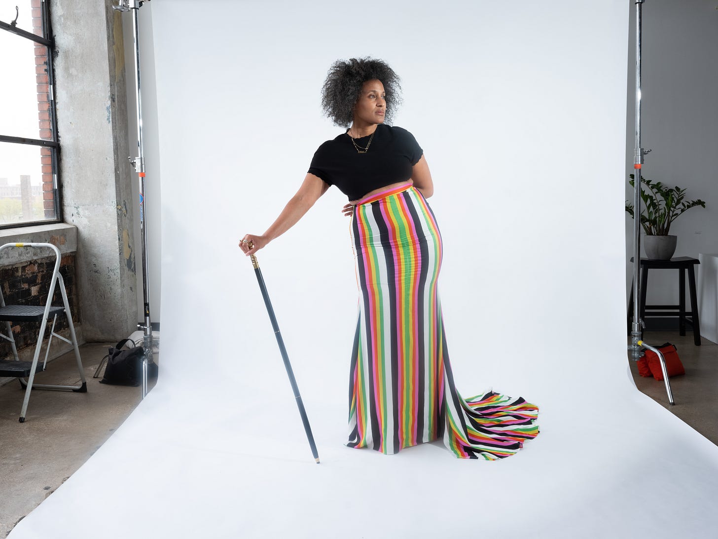 Dr. Cadet stands on a white backdrop in a studio wearing a stripped long skirt, black crop top and black cane with gold top. She looks to the side and her face says "I am amazing."