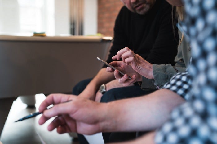 A woman using a phone and two people sitting next to her and taking notes. The person in back seems to be the facilitator, while the person in front seems to be taking notes with a pen. A woman using a phone and two people sitting next to her and taking notes. The person in back seems to be the facilitator, while the person in front seems to be taking notes with a pen.