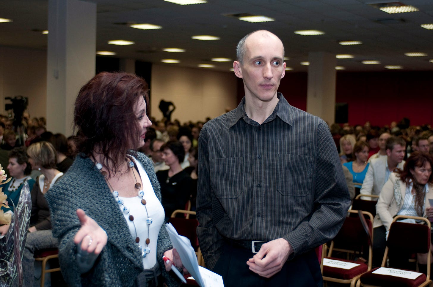 A rendezvény házigazdájával Manhertz József beszélget. A képen egy hölgy látható a bal oldalon, egy férfi a jobb oldalon. A hölgy beszél a férfi felé nézve, és jobb tenyerét a kamera felé mutat. A férfi jobbra néz, miközben a hölgyet hallgatja.