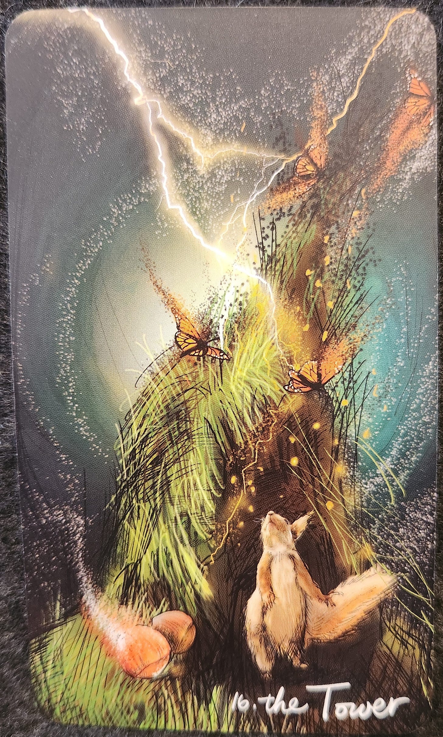 A tarot card labelled XVI Turm. A tall tower has been struck by lightning with flames pouring from the windows, and people falling to their death on the jagged rocks below. There is a large crown which has toppled from the top of the tower. The sky is dark and stormy, with clouds and golden rain. 
