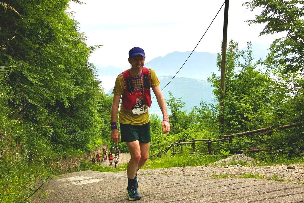 Ragazzo con pantaloncini, maglietta e cappellino da trail running mentre guarda in camera sorridente, salendo una cementata in salite circondata da alberi. Sullo sfondo compaiono delle montagne.