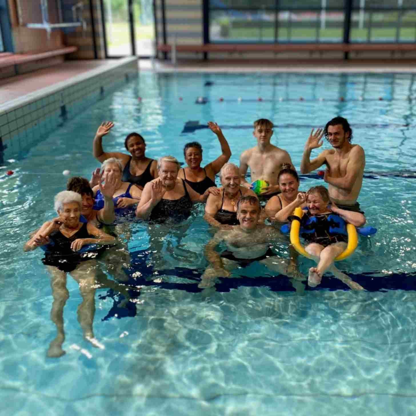 A group of people, about a dozen, wave hello from an indoor swimming pool. They are a mix of young and old. 