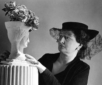 1) Constance is a white woman in a dark outfit and hat with lace coming off the back holding a sculpted vase shaped like a face with roses sticking out of the top, her expression curious and intrigued, against a plain backdrop. The vase sits on a ridged pillar, and she is examining the flower arrangement to see if she's happy with how it looks. She has on a small hoop earring, and is wearing rings on her left ring finger. 2) A black-and-white photo of a white woman in a head scarf arranging flowers, with a white man observing her work. The setting feels elegant and focused. Constance is arranging a floral bouquet, and she is holding a flower that she is about to place. She has on a pearl necklace, and earring, and she has rings on her right ring finger. She is wearing dark clothing, and the man watching behind her is wearing a suit.