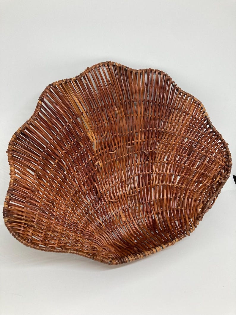 May include: A large, brown, woven wicker basket with a scalloped edge. The basket is made of thin, brown, woven strips of wicker. The basket is empty and is sitting on a white surface.