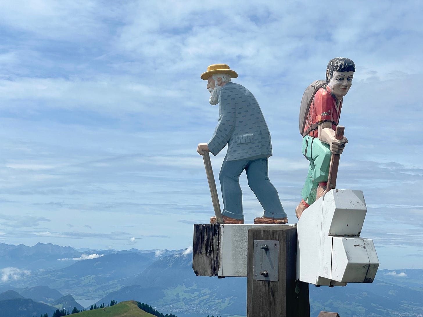 Carved figures of an old man and a young man on a sign. Mountains in the background.