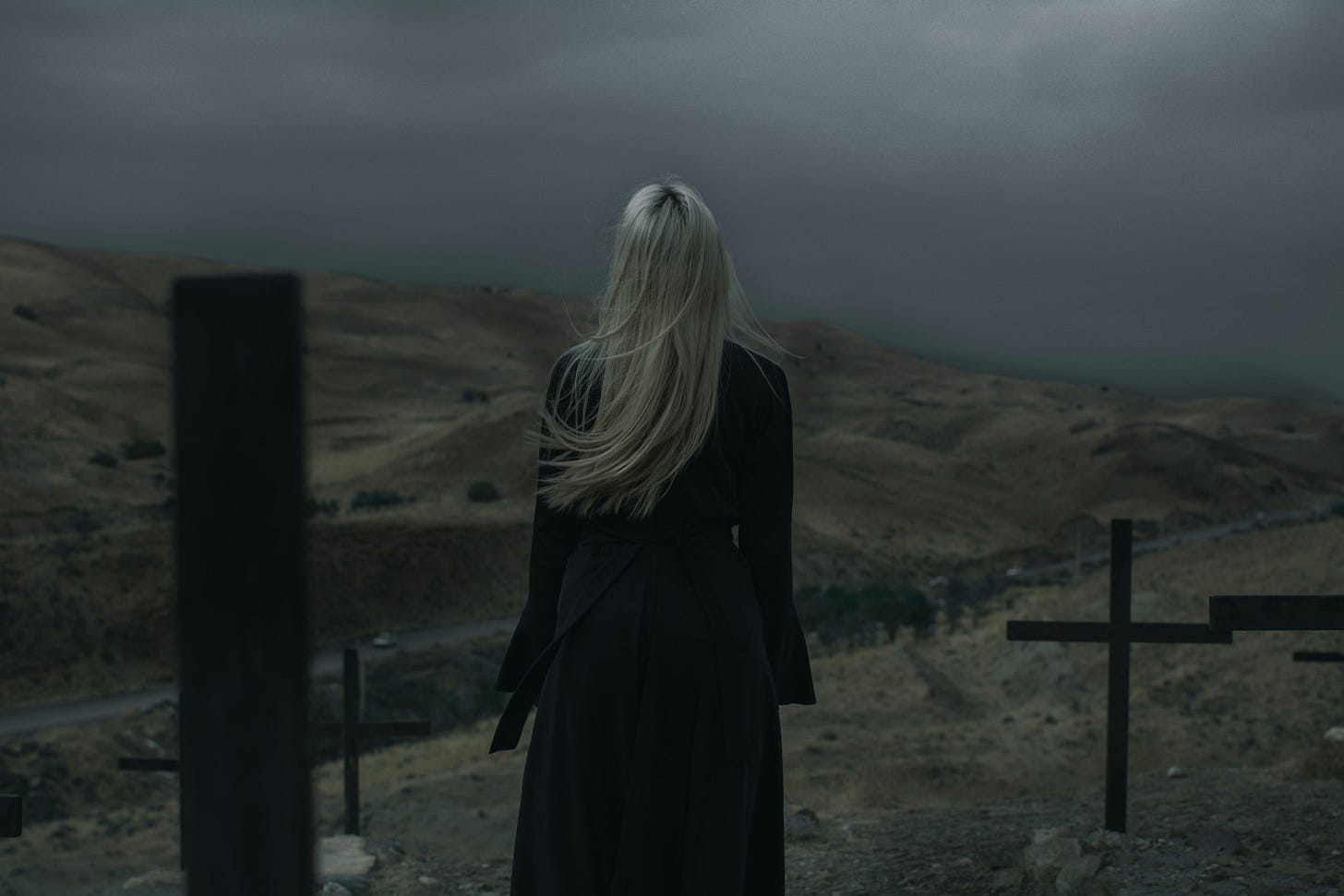 A woman with white hair in a black dress stands amongst wood crosses and looks out into a foggy horizon.