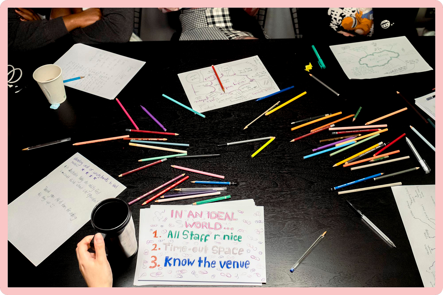 A photo of pens, colouring pencils and sketches, on a black wooden table.