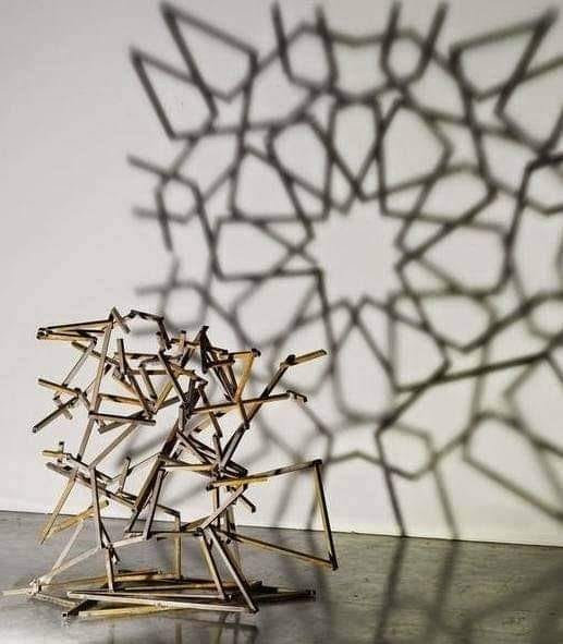 a chaotic sculpture that is being lit in such a way that it throws a shadow of an ordered, symmetrical pattern.