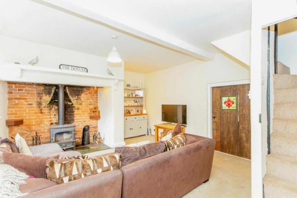 A living room with large sofa (too big for the room tbh), TV, and a door with stained glass window. There's a multifuel burner in a brick inglenook