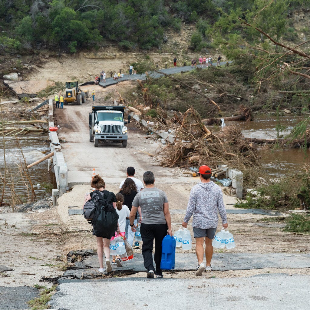 A family carries supplies toward a damaged bridge that’s surrounded by fallen trees. 