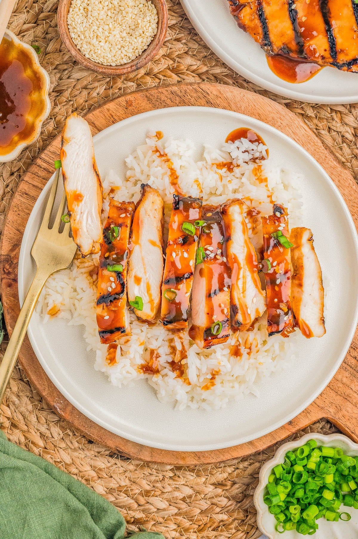 A plate of sliced grilled chicken with sauce, served over white rice and garnished with chopped green onions. A fork is placed on the side of the plate.