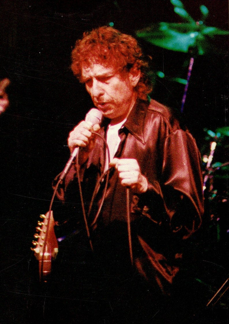 May include: A person singing into a microphone while wearing a brown leather jacket and a white shirt. May include: A person singing into a microphone while wearing a brown leather jacket and a white shirt.